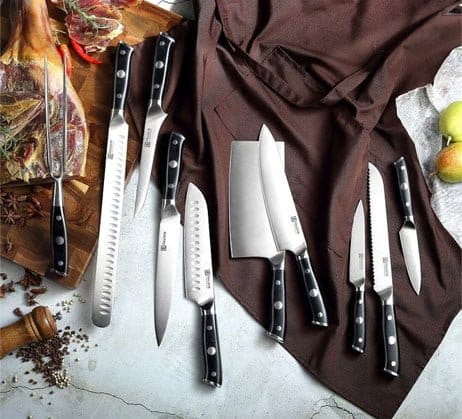 A collection of various chef's knives, including a cleaver and bread knife, displayed artistically on a kitchen countertop with herbs, spices, and a brown cloth.